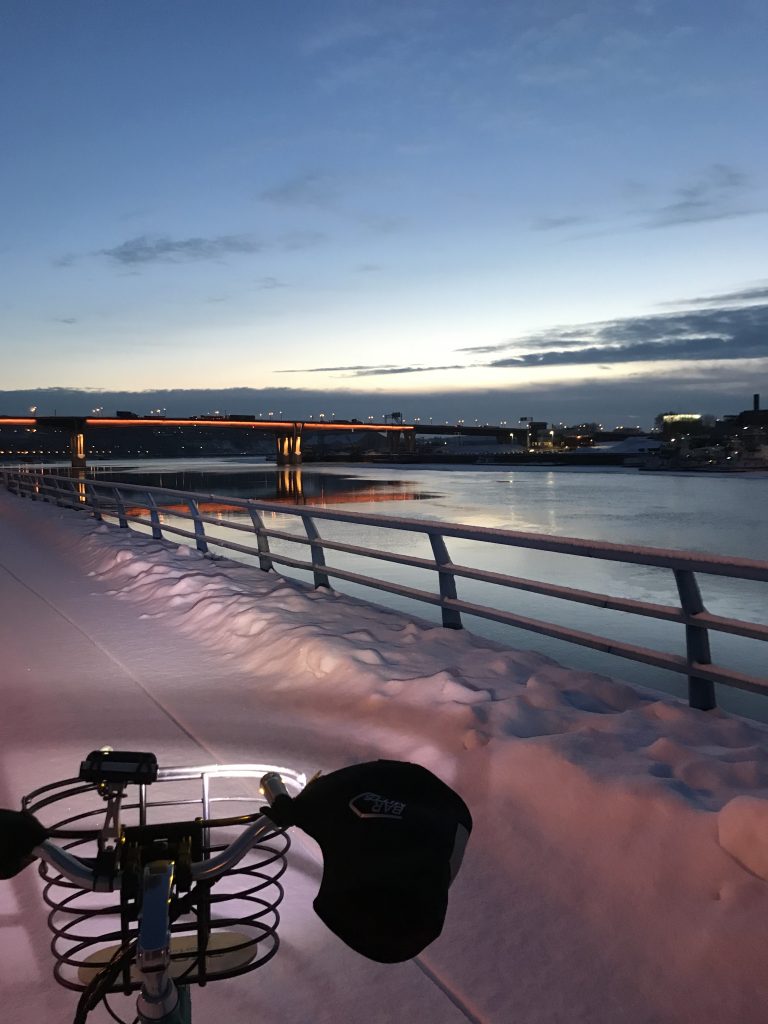 Snow covered bike path with bike basket and bar mitts in foreground. River with railing and bridge, plus dawning sun and grey clouds.