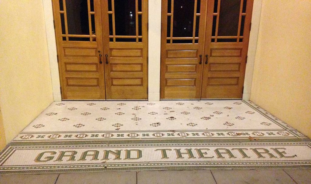 Grand Theatre in gray-green tile with gray-green and gold patterns on off-white backgorund, honey-gold wooden doors in background