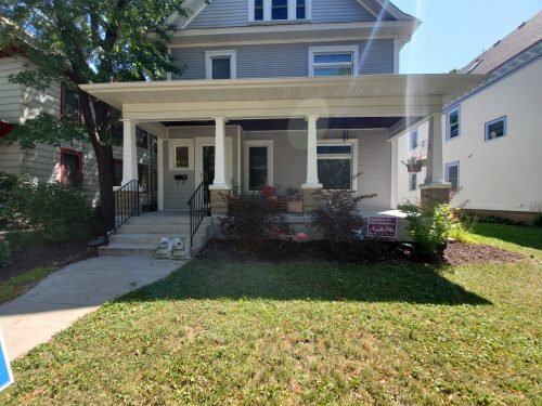 Front of a house with blue siding and white trim, large front porch with seating and potted flowers