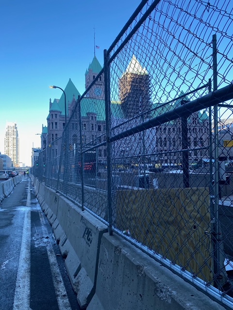 Concrete barrier topped by a fence in the foreground, lining a path of the road blocked off for pedestrians. In the background behind the fence is Minneapolis City Hall, which is brown brick and topped by a green roof with spires and towers.