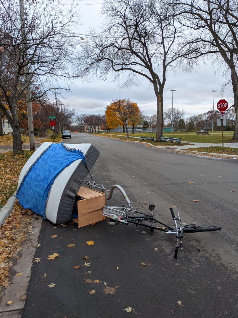 The trailer and bike and mattress are on their side in a gutter on a quiet street in Saint Paul on a cloudy November afternoon.