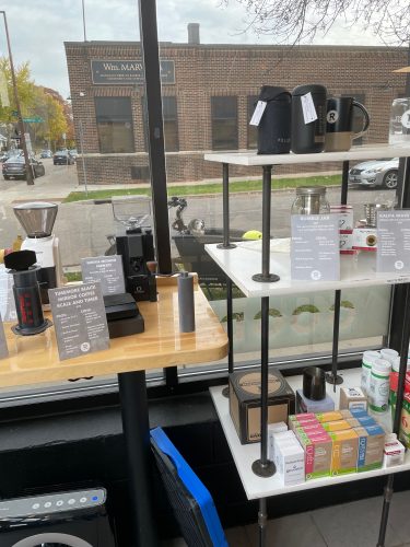 A dog sitting in a cargo bike outside is seen beyond the inside shelves of coffee tools for sale at a coffee shop.