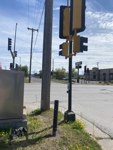 A pedestrian call button on a traffic light pole that's set back from the sidewalk, with another utility pole blocking access.