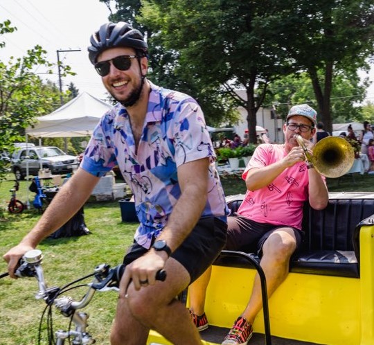 A man on a bike hauling a man playing a tuba in a bike cart.