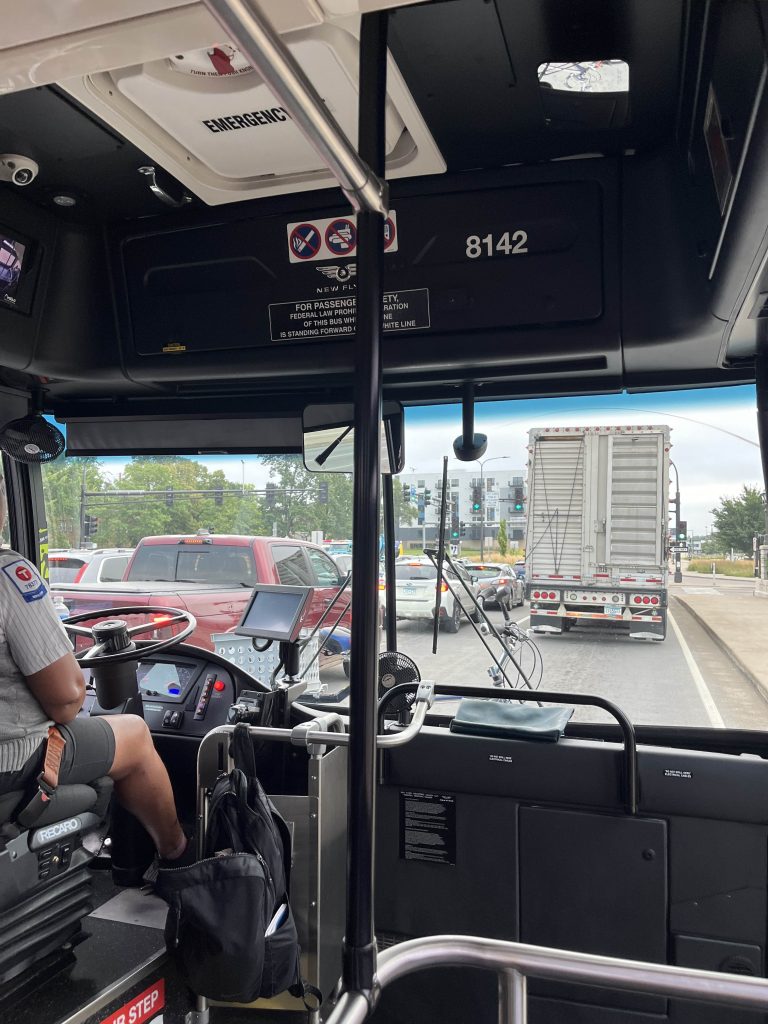 View looking out the front of a bus headed to the Minnesota State Fair. The road is busy with cars and trucks.
