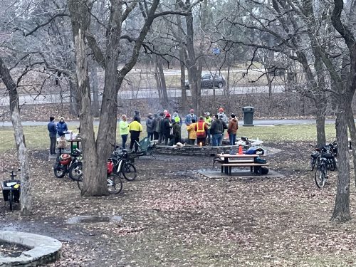People standing by fire in park. Many bikes visible.
