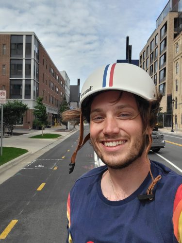 A smiling man wearing a white helmet with red and blue stripes, posing for a selfie on the Second Street bikeway. The background features modern apartment buildings, a bike lane with yellow markings, and a partly cloudy sky.