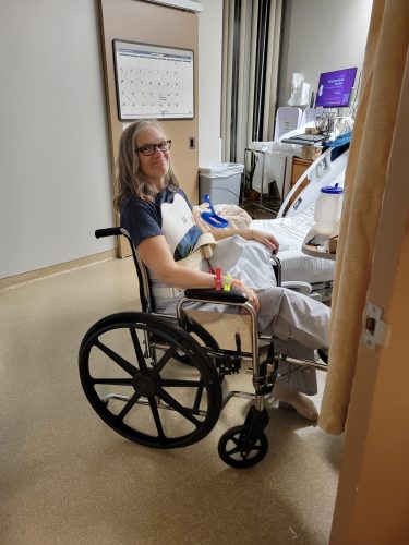 Julia Klatt Singer, a middle-aged woman, is shown smiling in a wheelchair in a hospital, with a hospital bed in the background.