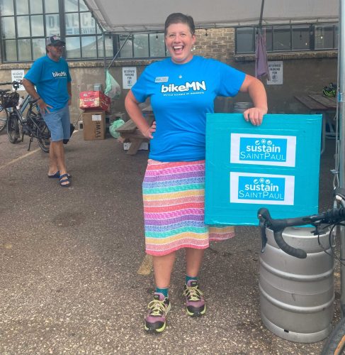 A laughing woman holds up a Sustain Saint Paul sign outside a brewery.