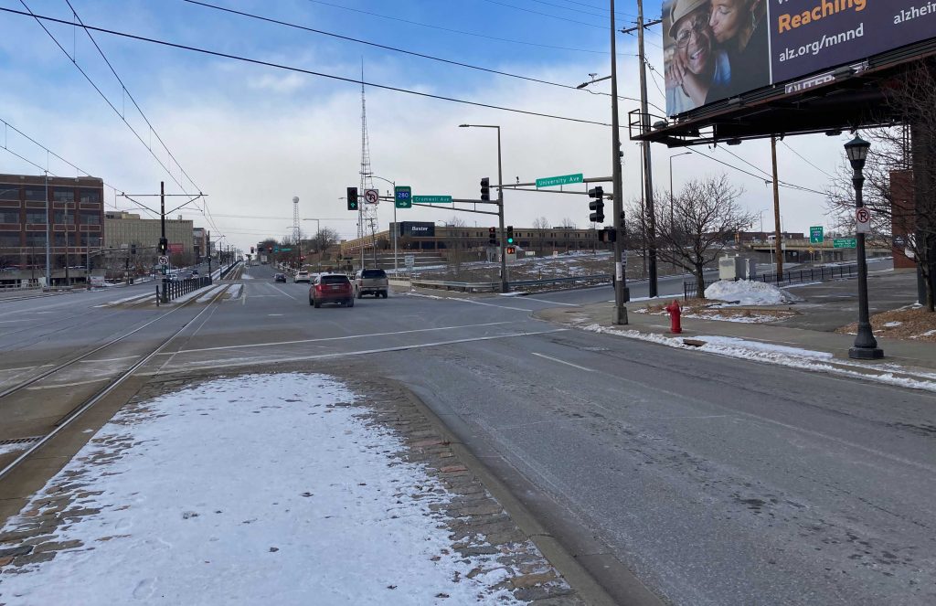 A wide urban intersection on a cold winter day, with light snow covering the sidewalks and patches of the road. The traffic lights and street signs mark the crossing of University Avenue and Cromwell Avenue, with cars driving toward the horizon.
