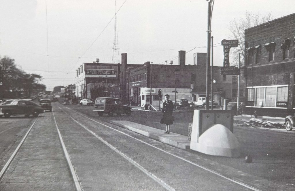 1940s image of University Avenue. Woman waiting for streetcar in middle of street, brick commercial buildings in background, cars turning.