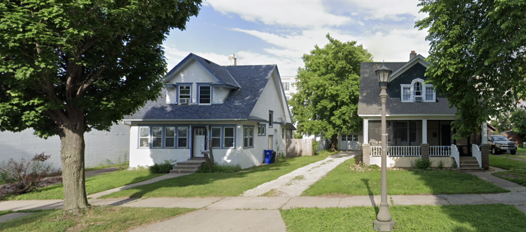 Two neighboring early 20th-century bungalows with steep gable roofs, dormer windows, and front porches sit side by side on a tree-lined suburban street. The house on the left is white with a blue roof and enclosed porch, while the house on the right is dark green with a covered porch and white trim. A vintage-style street lamp stands near the sidewalk, and a gravel driveway separates the two homes.