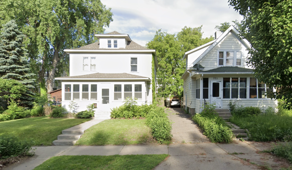 Two older homes sit side by side on a residential street, surrounded by greenery. The house on the left is a white American Foursquare with a symmetrical facade, a dormer window, and a large enclosed front porch. The house on the right is a traditional farmhouse-style home with pale yellow siding, a gable roof, and an enclosed front porch with black trim around the door. A narrow driveway runs alongside the right house, leading to a parked car in the background.