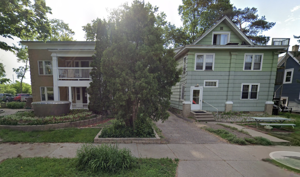 Two multi-unit residential buildings stand side by side on a tree-lined street. The building on the left is a two-story brick structure with white columns and a second-floor balcony, featuring a landscaped front yard with a curved planter. The building on the right is a large, pale green wooden duplex with a gabled roof, multiple windows, and an external staircase leading to an upper-level entrance. A picnic table sits in the yard, and a driveway runs between the two buildings.