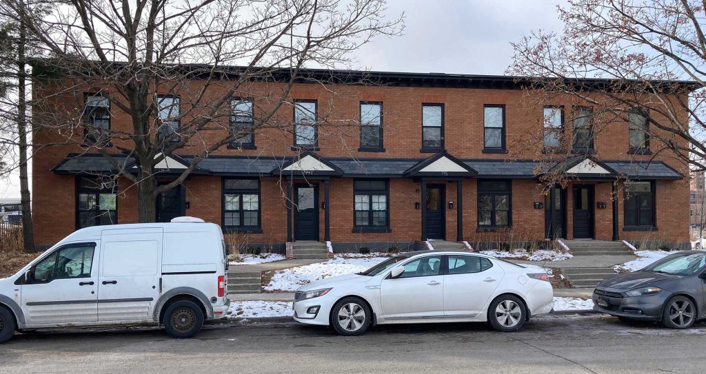 A row of modernized brick townhomes with black trim and awnings sits on a residential street with a light dusting of snow on the ground. Each unit has a black front door with a small gabled awning above, along with large rectangular windows.