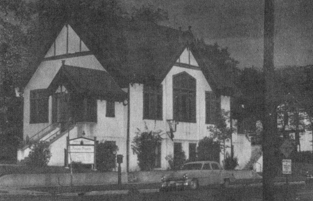 A historic black-and-white photograph of a Tudor-style church with steep gable roofs, decorative half-timbering, and large arched windows. The entrance is elevated, with a staircase leading up to a covered doorway. A small sign stands near the sidewalk, and vintage cars are parked along the street.