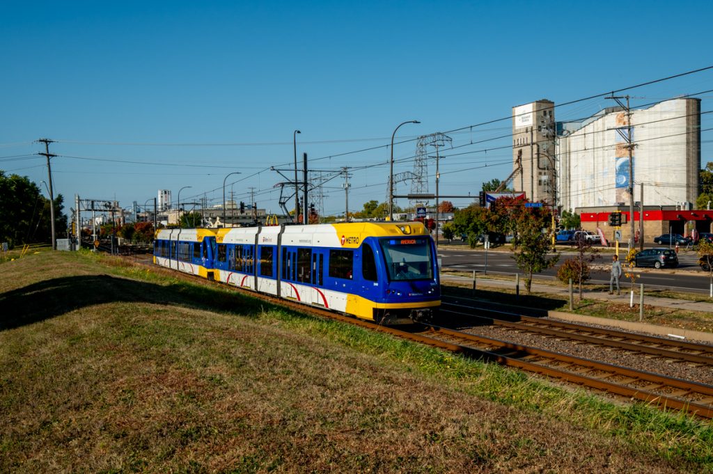 A Blue Line train with a grassy strip of land in the foreground and grain elevators across Hiawatha Avenue in the background.