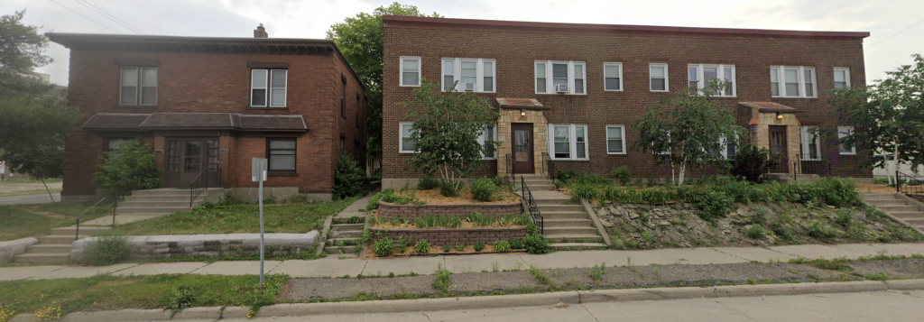 A pair of old brick apartment buildings sit side by side along a slightly elevated lot with stairs leading up from the sidewalk. The left building is a two-story structure with a flat roof and a small overhang above the entrances. The right building is a longer, two-story row-style apartment with multiple entrances, each featuring small awnings and steps leading to the doors.