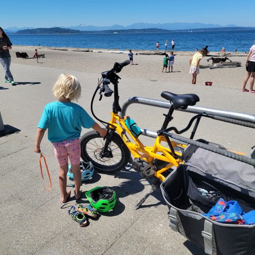 A child with an e-bike, helmets and gear at a sunny beach.