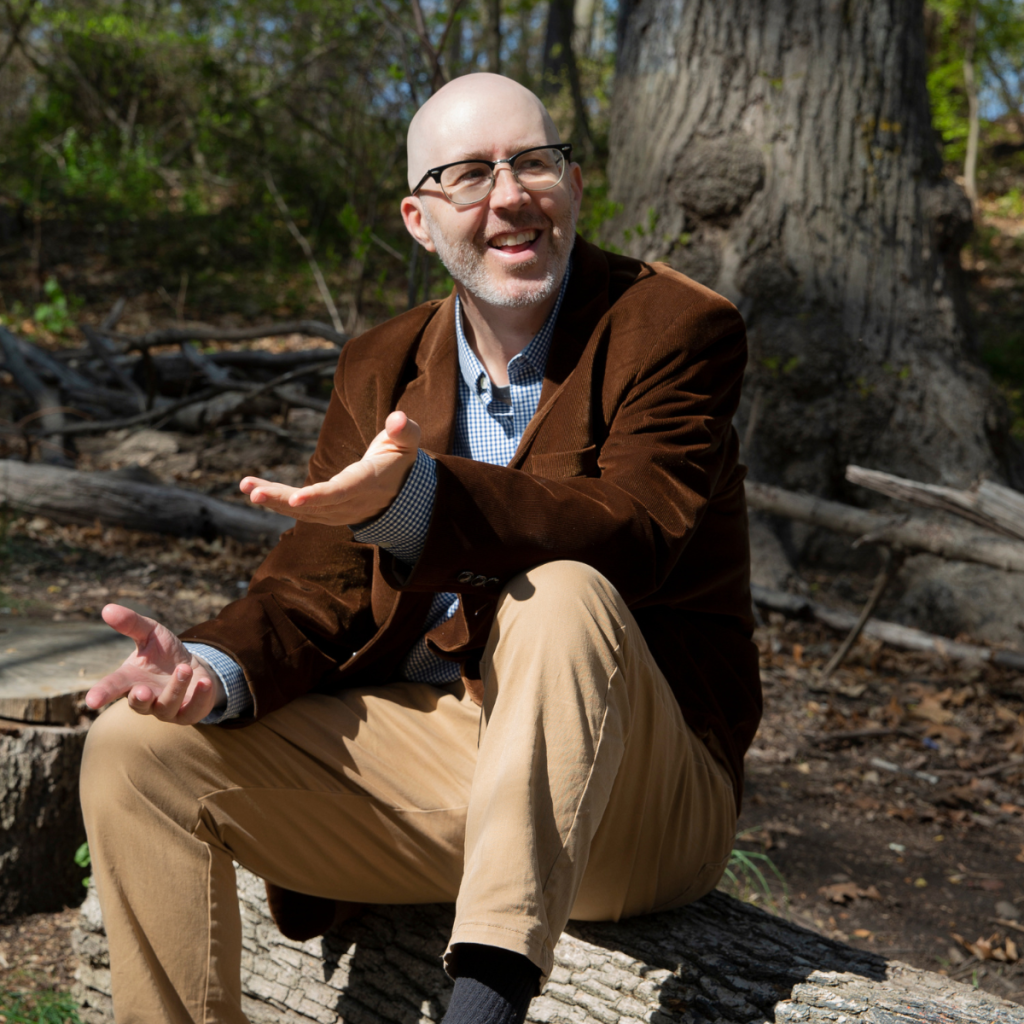 A man sits on a log in the woods, gesturing toward the camera.