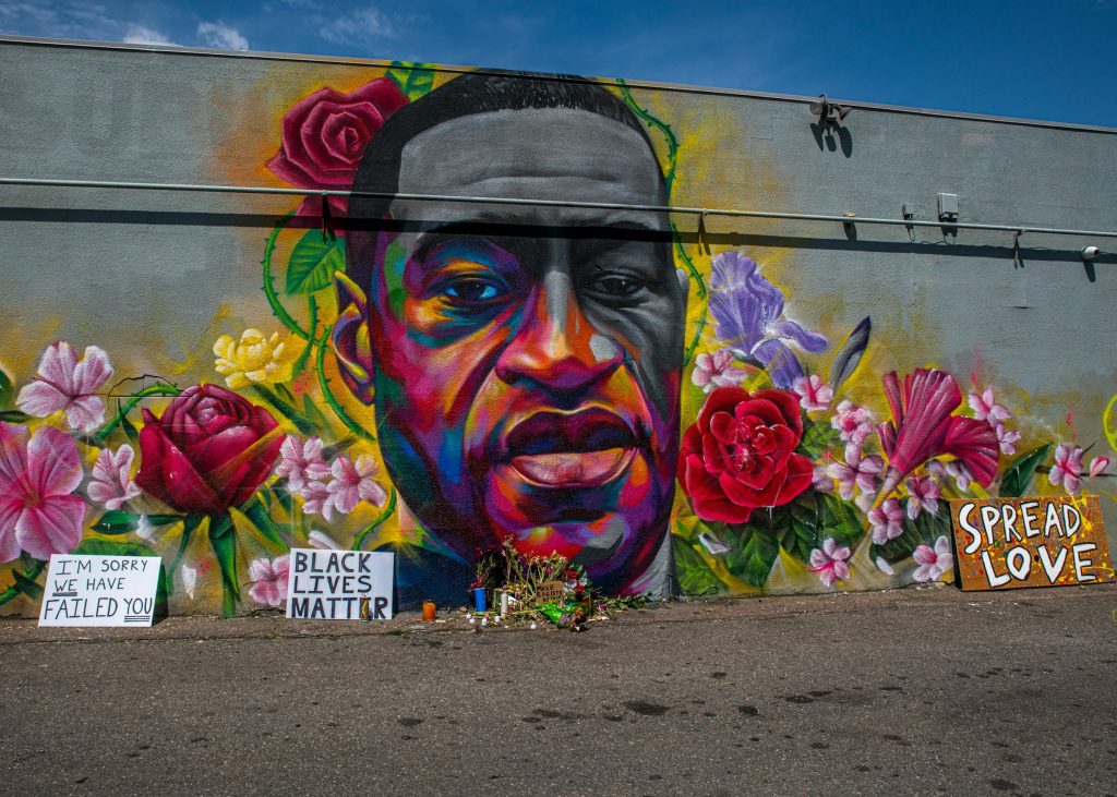 An art installation on a concrete wall shows the head of George Floyd, flanked by colorful flowers and signs such as "Black Lives Matter."
