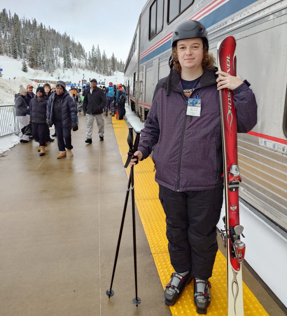 Board member Ian Gaida stands with his downhill skis and poles on a train platform with hills and trees in the background.
