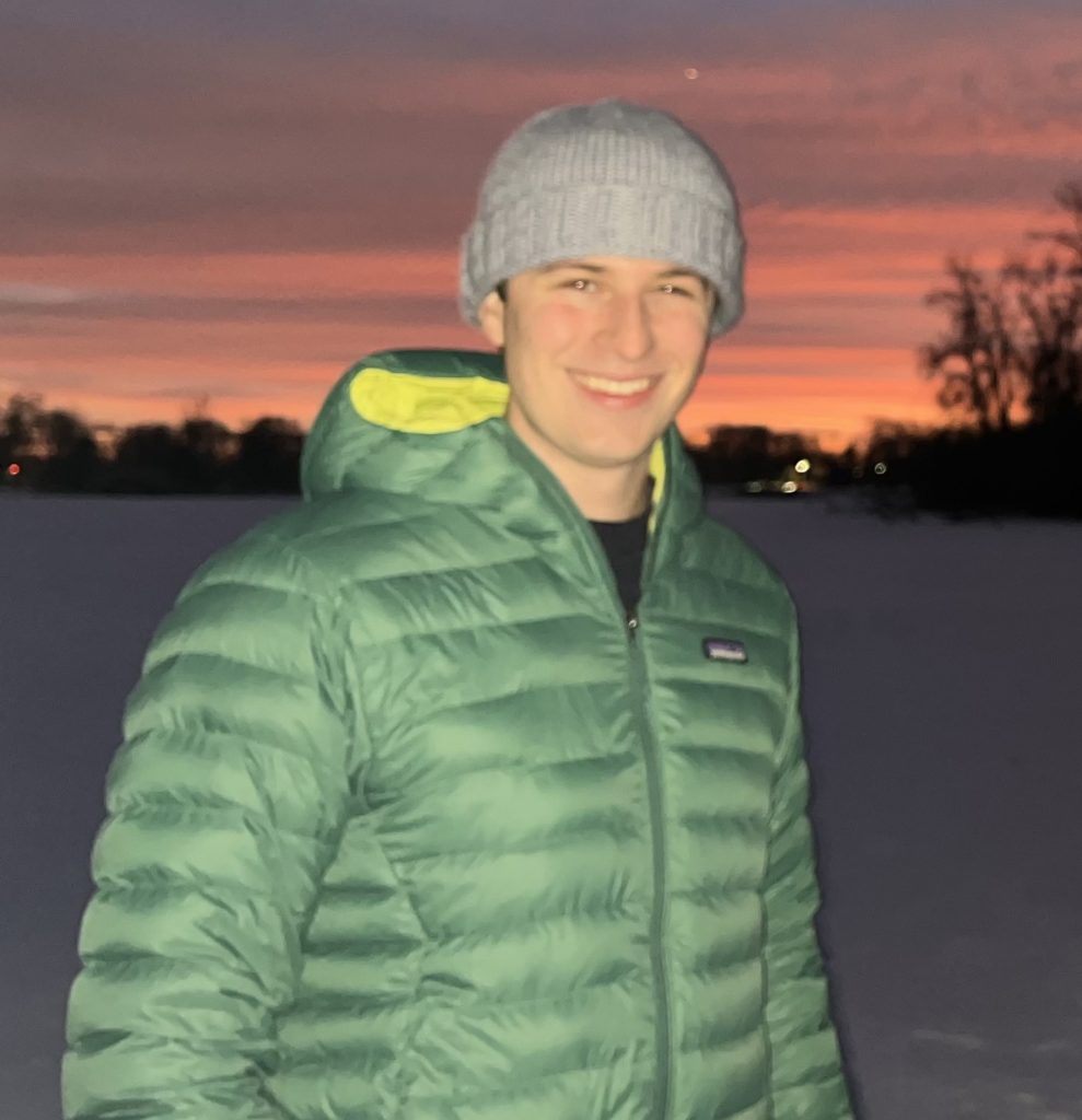 A man wearing a hat and winter jacket poses by a lake against the sunset.