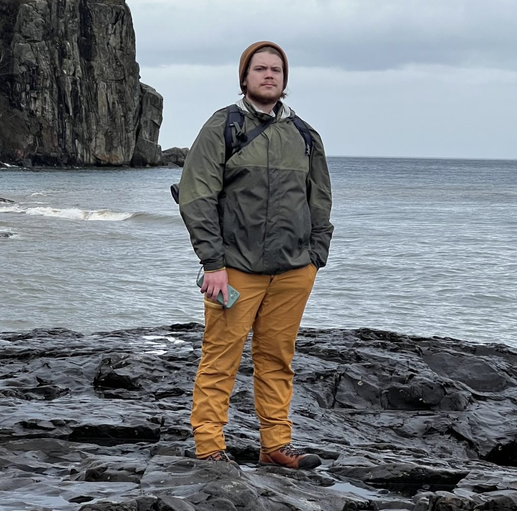 A full-body shot of a man standing on gray rocks with his back to Lake Superior.