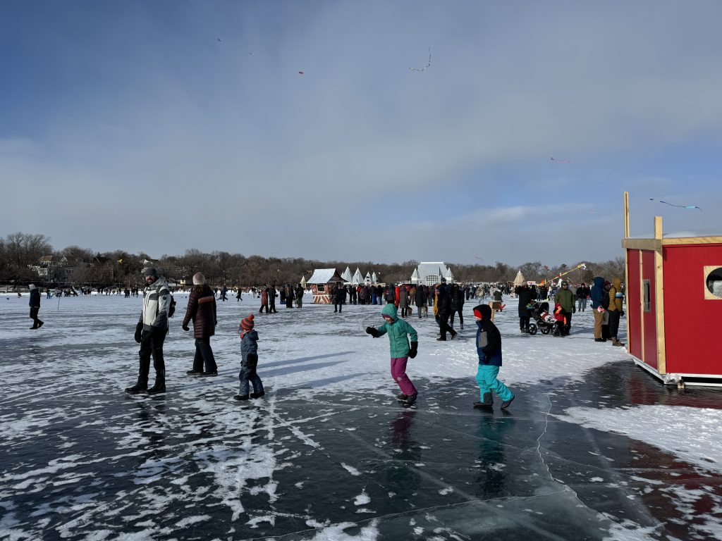 Lake Harriet hosts the Art Shanty Project every January, shown here with adults and children walking on the frozen lake amid small shanty structures.