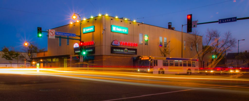 Cars speed through the intersection at Lake & Bloomington Avenues at night. A bus waits for the light to change. 