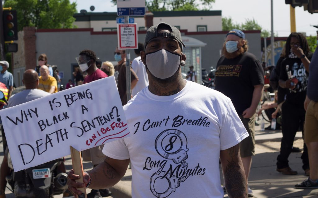 A Black man in a white t-shirt that reads "I Can't Breathe 8 Long Minutes", and holding a sign that reads "Why is being Black a death sentence" stands at a protest in George Floyd Square. 