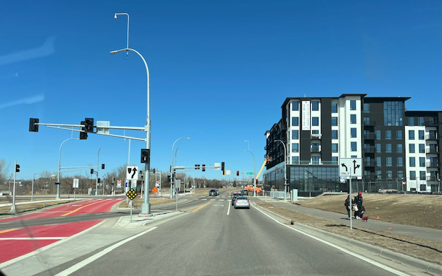 View approaching Helmo Station, where the METRO Gold Line busway transitions from the center median to the west side of the street. A multi-story apartment building lines the right side, with red-colored bus-only lanes and multiple traffic signals guiding vehicles through the intersection.