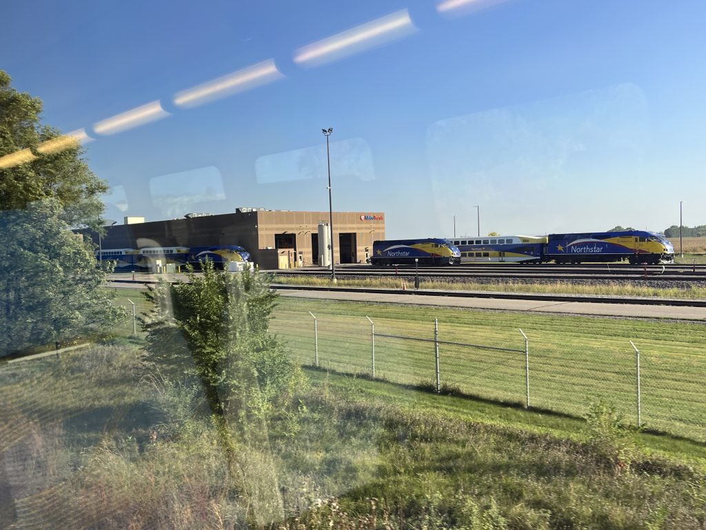 Three Northstar commuter trains are parked on parallel tracks outside a tan maintenance facility in Big Lake, viewed through the window of another train.