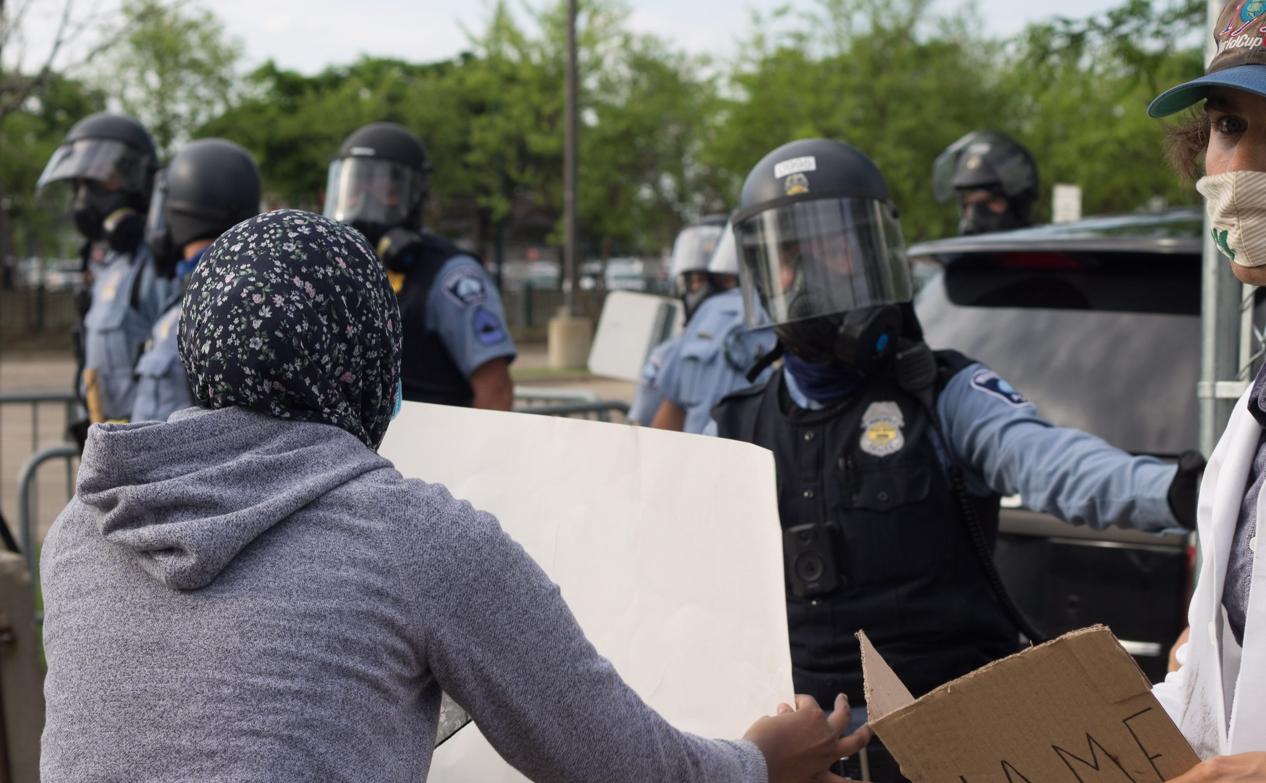 A woman in a hijab holds a sign up to a police officer in riot gear,