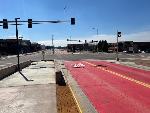 Looking south from Tamarack Station along Bielenberg Drive, with a dedicated red bus-only lane in the center and traffic lights ahead. Sidewalk infrastructure is present, but the commercial developments on both sides of the road show minimal pedestrian access or connectivity.