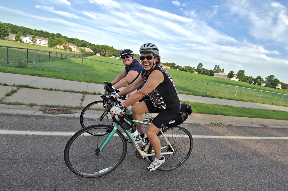 Two cyclists on a road with fields and a housing subdivision in the background.