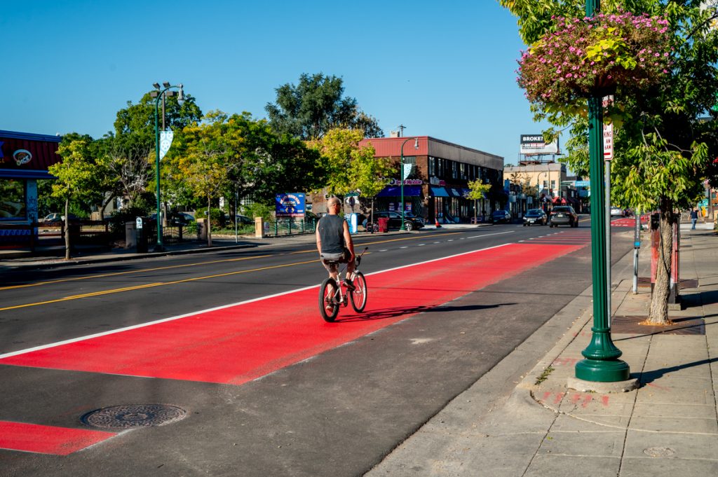 A cyclist rides in a red lane on a city street, their hands at their sides. There are trees and buildings in the background.