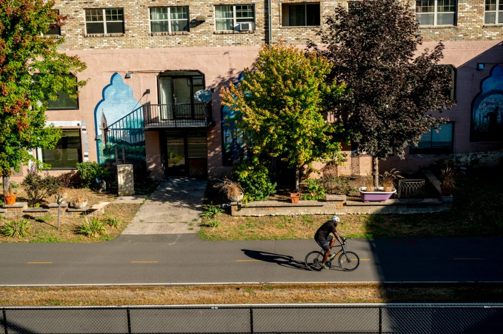 A cyclist in black rides on the Midtown Greenway bike path with a building and trees in the background.