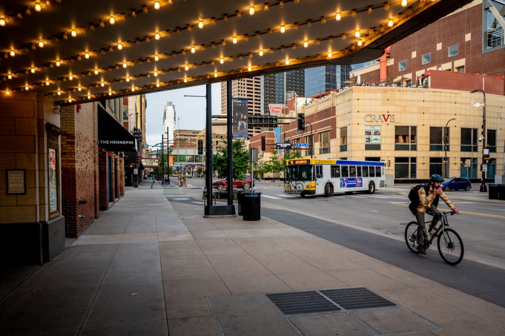 A cyclist rides a bicycle in the foreground aside a sidewalk. There is a bus and buildings in the background.
