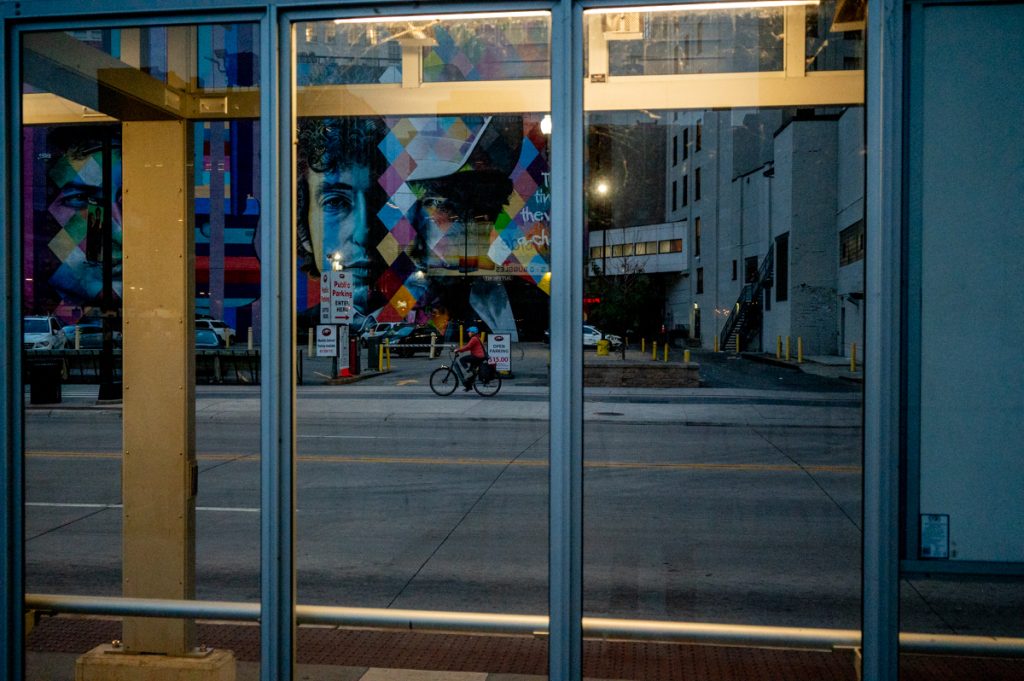 In the reflections of a buildings glass front, the photo depicts a cyclist on a street with a colorful mural above them.