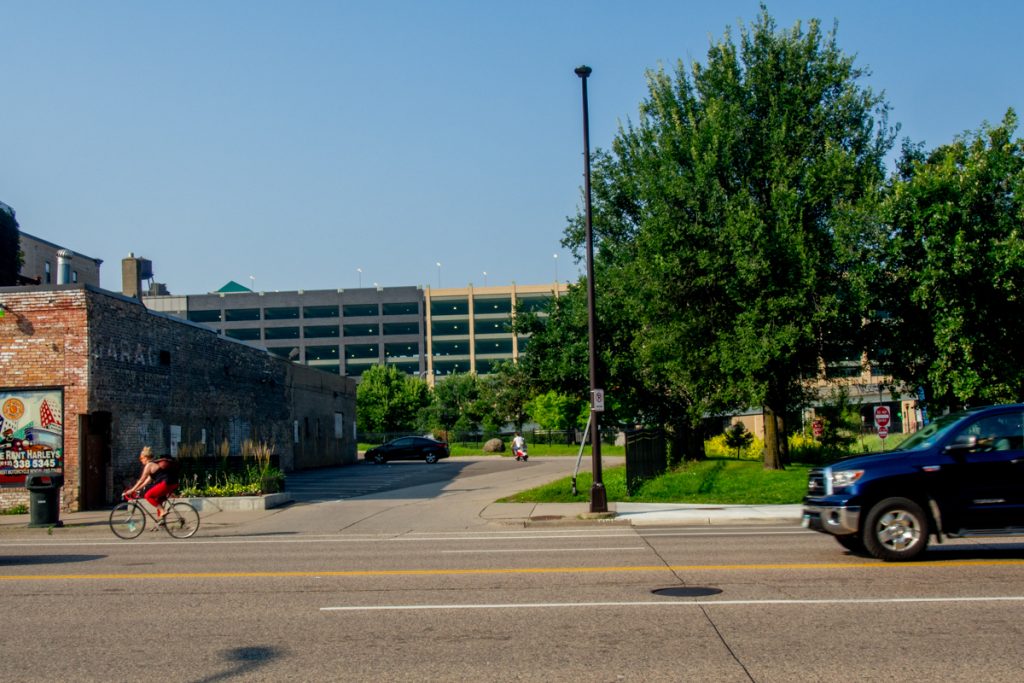 A city street photo shows a cyclist and a vehicle with trees and buildings beyond them.