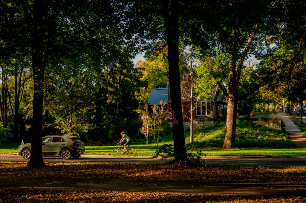 A lone cyclist on a residential street in Minneapolis, as seen from a distance through some trees.