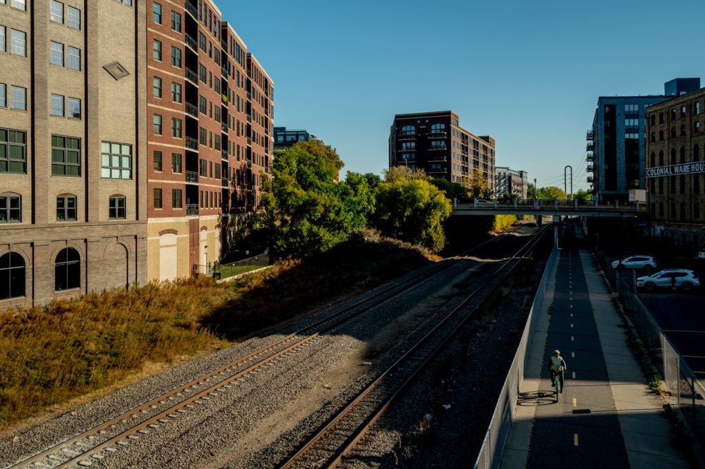 A cyclist rides on a bike trail on the right side of the photograph with train tracks besides them. There is shadow over the trail and buildings and trees in the background.