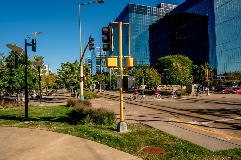 Street scene with a group of cyclists in an intersection in the center. There are grass, trees, and buildings throughout the photo.