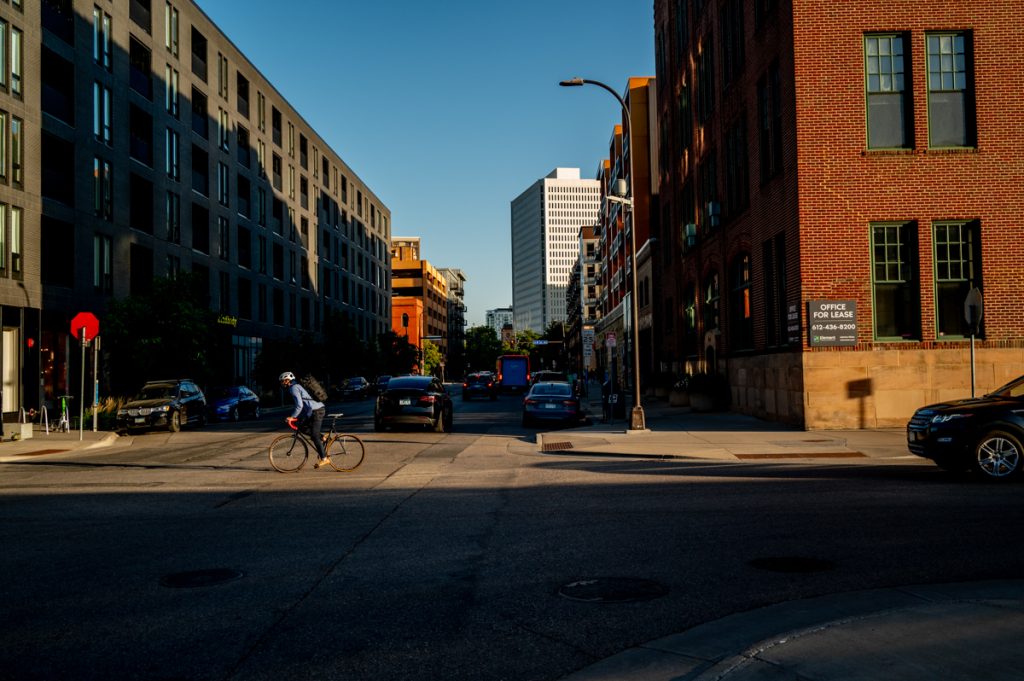 A city street is cast in shadow with a cyclist riding in a sliver of sunlight. There are cars and buildings in the background with the street in the foreground.