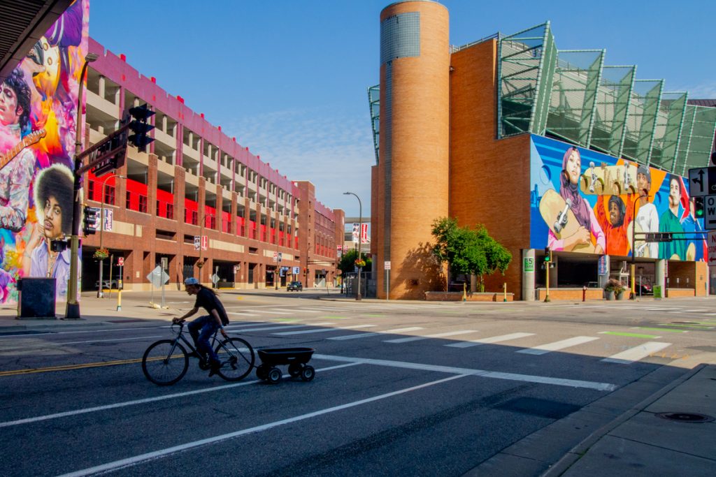 A cyclist, cast in shadow, pulls a wagon from their bike. Colorful murals depicting various people adorn buildings in the background.