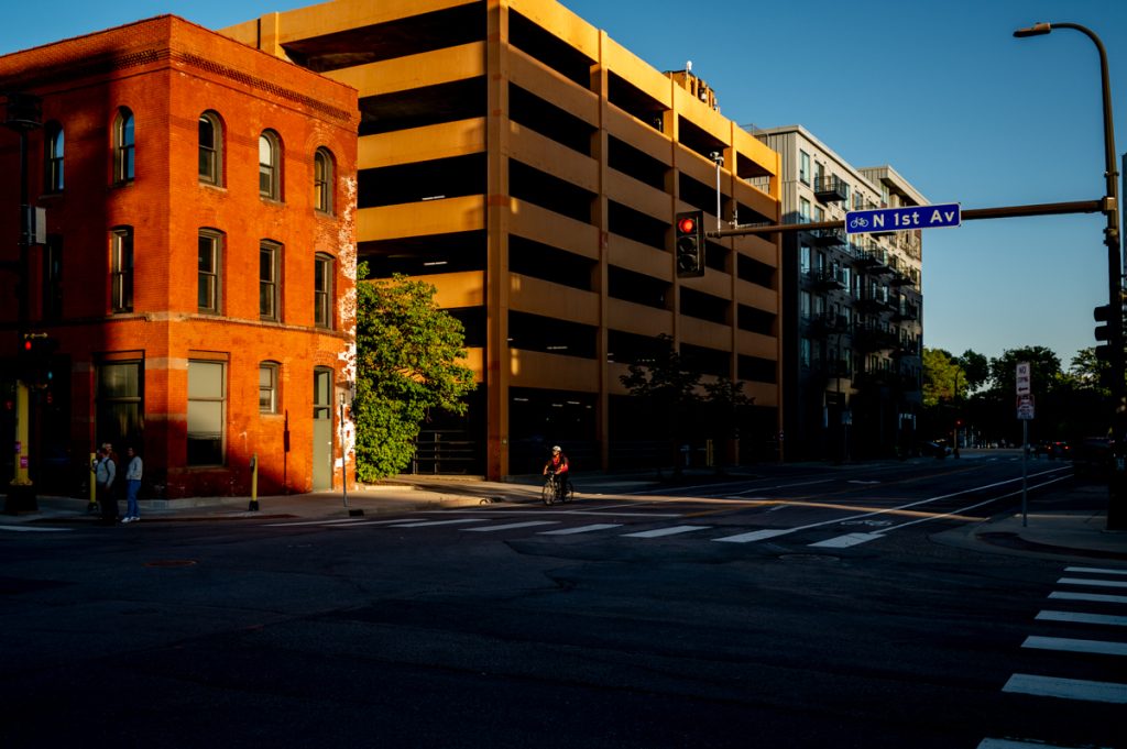 A shadowy photo shows darkness cast over buildings, a city street, and a cyclist. 