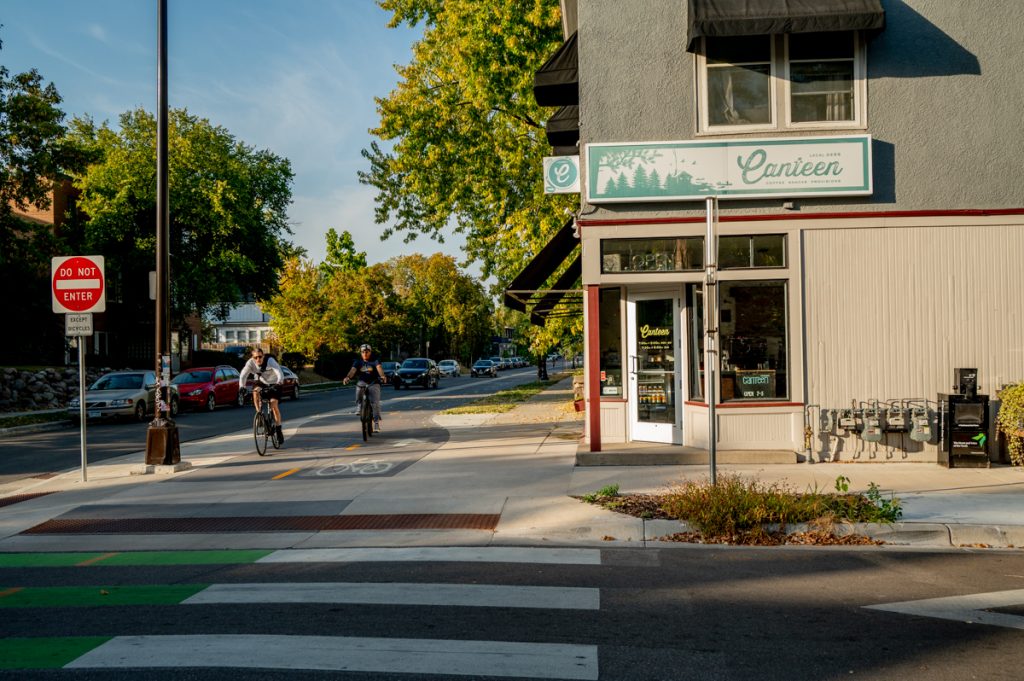 Cyclists ride on a separated bicycle path aside a business toward an intersection. 