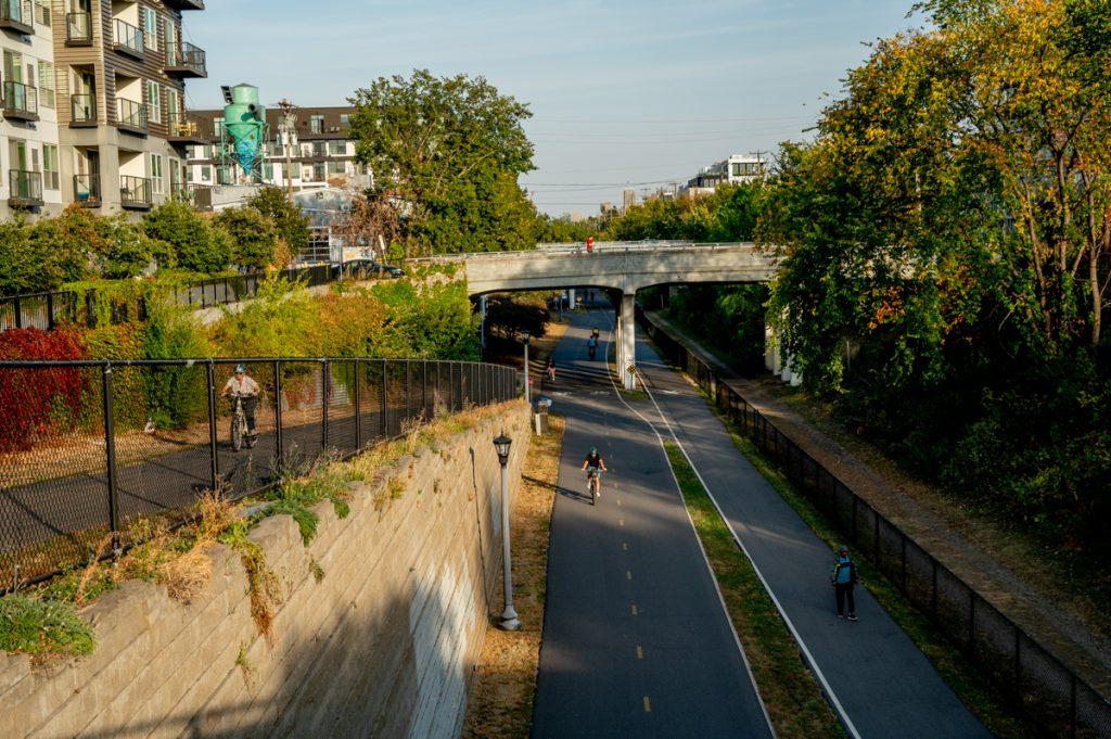 Shot from above, there is a shot of cyclists on a bike path and a ramp to the (not pictured) street. There are trees and buildings in the background.