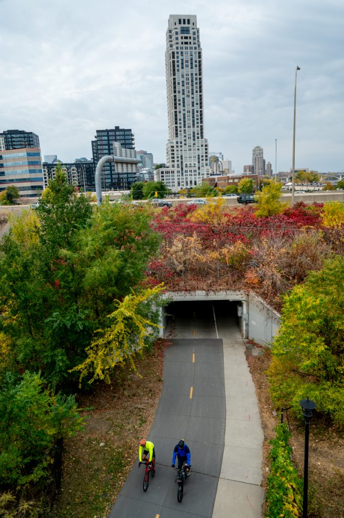 Two riders are shown from overhead on a bike trail. They are surrounded by grass and trees. The opening to a tunnel is behind them with a shot of the Minneapolis skyline beyond that.